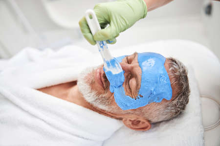 Cropped photo of a spa staffmember applying blue foam to an elderly man face with a spatelの写真素材