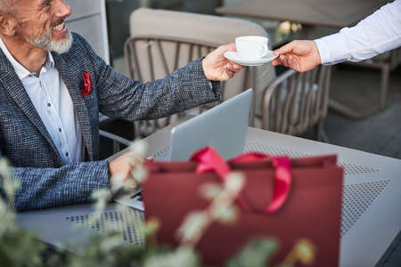 Cropped photo of a mirthful senior man taking a cup with a hot beverage from a waiter at the cafeの写真素材