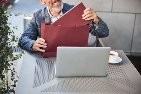 Contented elderly man taking a generic red box out of a red gift bag while sitting at a cafe tableの写真素材
