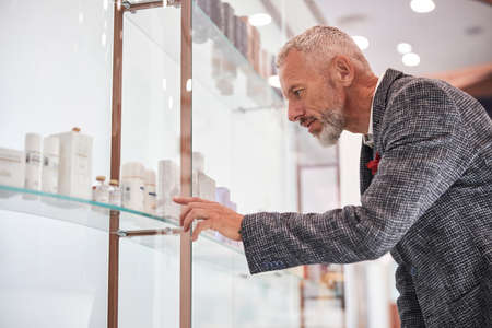 Thoughtful aging gentleman pointing to a a cosmetics bottle on a shelve during shoppingの写真素材