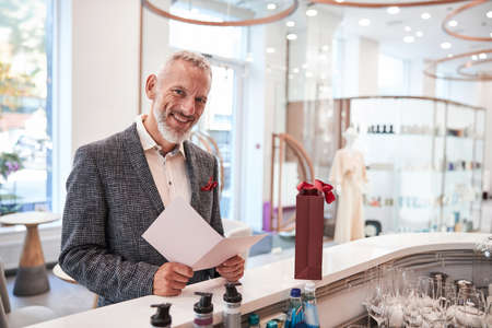 Cheerful elderly man in an elegant blazer holding a white paper and smiling at the cameraの写真素材