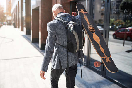 Back view of a senior citizen with a backpack carrying his longboard and walking