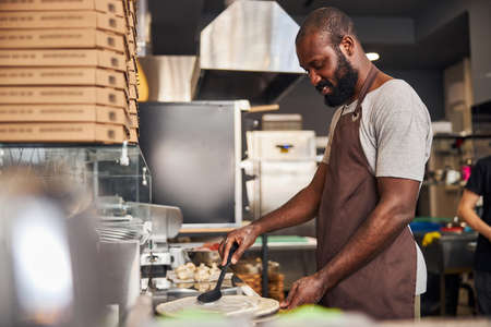 Young bearded male is standing at table and spreading sauce on dough with spoon in kitchen