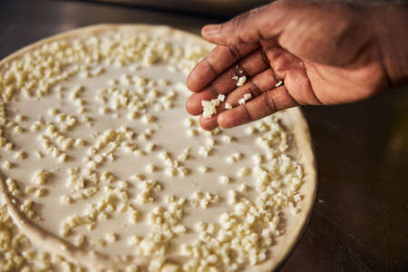 Top view cropped head close up of man hand dropping grated cheese on dough on table