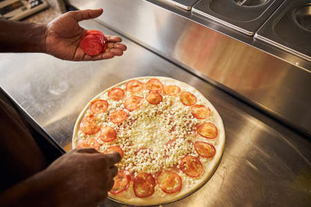 Top view cropped head close up of male in apron cooking and putting tomato slices on doughの写真素材