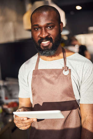 Waist up portrait of smiling bearded handsome man in apron standing in professional kitchen and holding tabletの写真素材