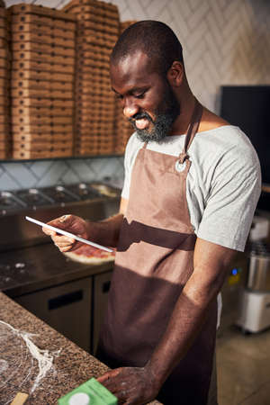 Happy bearded handsome man in apron is standing in restaurant kitchen and having fun with tabletの写真素材