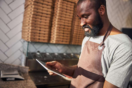 Cheerful young bearded male is making pizza in professional kitchen and using internet on touchpad