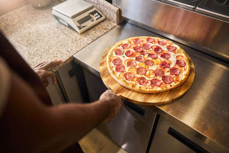 Close up of male hand holding wooden board with freshly baked pizza with salamiの写真素材