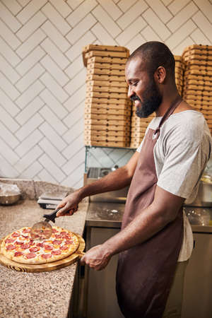 Handsome Afro American man in apron slicing pizza with round cutter wheel and smilingの写真素材