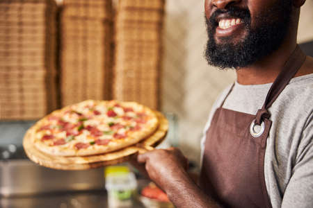 Close up cheerful male chef in apron holding wooden board with delicious pizzaの写真素材