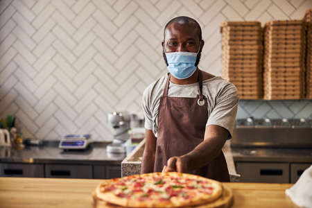 Handsome male worker in protective face mask standing at the counter with freshly baked pizza in pizzeriaの写真素材