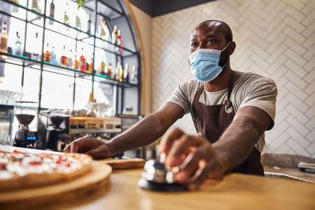 Afro American man in protective face mask using call bell while standing at the counter with freshly baked pizzaの写真素材