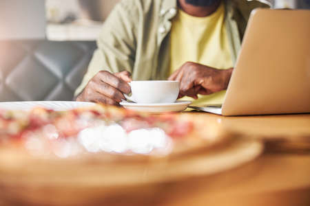 Close up of male freelancer holding cup of hot drink while sitting at the table with modern laptop...