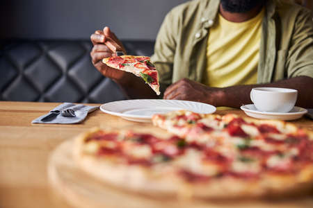 Close up of male hand holding slice of pepperoni pizza while gentleman sitting at the table with cup of coffee and white plateの写真素材