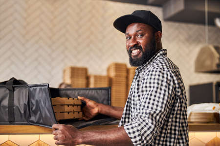 Handsome male courier looking at camera and smiling while packing cardboard pizza boxes in insulated...