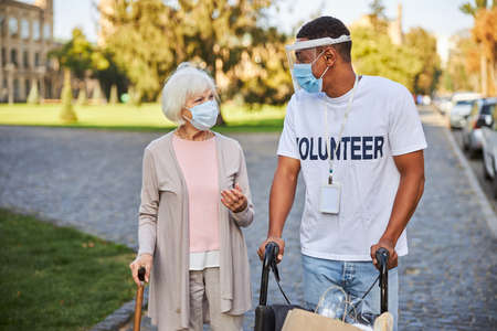 Young man in protective shield pushing a wheelchair and looking at the elderly woman with a walking stick by his side outdoorsの写真素材