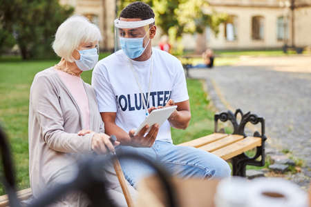 Young male volunteer wearing a protective shield while sitting on a bench with an aged woman in medical mask and showing a modern tabletの写真素材