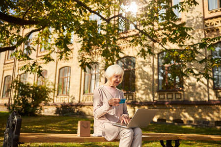 Senior woman with grey hair looking at her bankcard in front of a laptop while sitting near the hospital buildingの写真素材