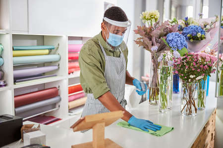 Young man in protective mask and gloves is cleaning surfaces with sanitizer at florist during pandemicの写真素材