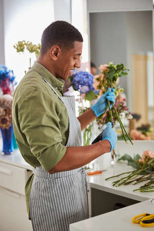 Cheerful young man is working in flower shop and using scissors for cutting green stemsの写真素材
