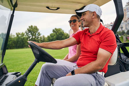 Joyful young Caucasian woman riding in a golf car with a smiling middle-aged male instructorの写真素材