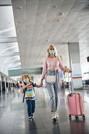 Mom keeping her son by the hand and going with her belongings at the airport gateの写真素材