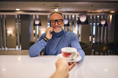 Merry bearded male in a jumper taking a cup of coffee from the barista hands