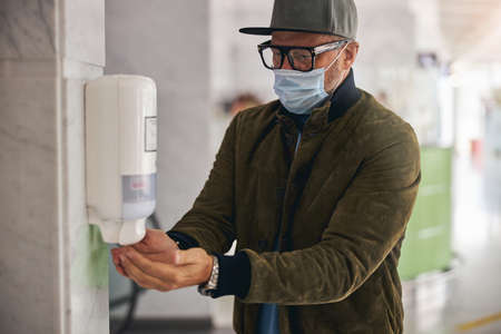 Serious man in a cap and a disposable protective mask using the touchless wall-hanging sanitizerの写真素材