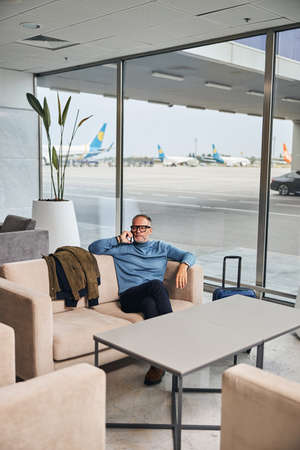Calm middle-aged male tourist with his cellphone sitting on the sofa in the waiting areaの写真素材