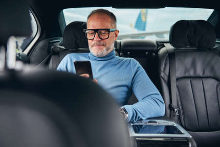 Front view of a smiling businessman reading news on his cellular phone in the cab