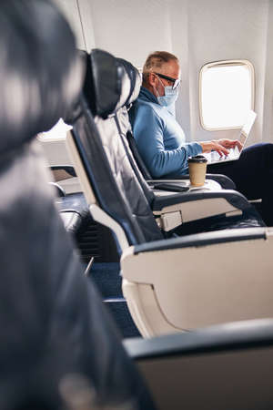 Side view of a man in eyeglasses and a protective mask typing on his laptopの写真素材