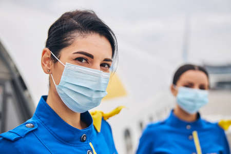 Portrait of a dark-haired young Caucasian flight attendant and her female colleague wearing medical...