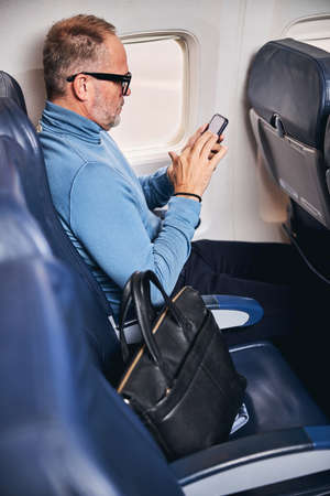 Side view of a focused man in eyeglasses using his mobile phone during the flight