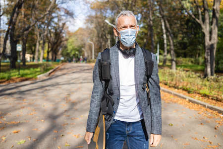Smiling trendy male in mask and earphones is enjoying free time in nature and going skateboardingの写真素材