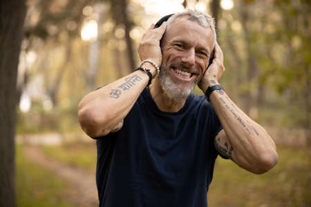 Portrait of smiling mature bearded guy listening to music in headphones while running in forest