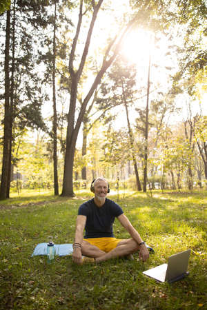 Man sitting in lotus position and using earphones and laptop for training in wood