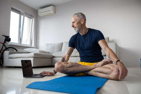 Full-length photo of a sporting man having a look at a laptop screen while sitting in a yoga pose