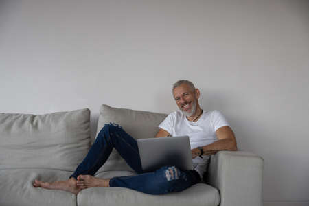 Satisfied man in torn blue jeans and white T-shirt lying in a couch corner with hands on a computer...