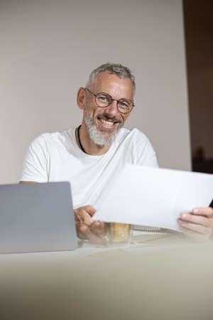 Close-up photo of joyful businessman picking up a paper and tea cup from the table while sitting at...