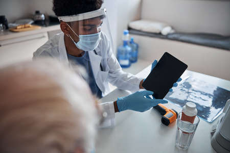 Young healthcare professional in a face shield holding the tablet computer in front of his patient