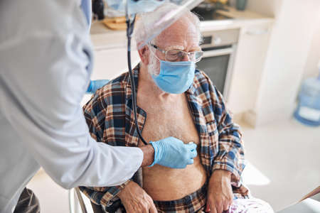 Doctor in a face shield and latex gloves examining the elderly man with a stethoscopeの写真素材