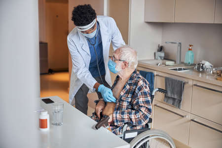 Focused African American healthcare professional listening to the senior male patient heartbeat...