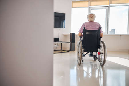 Back view of a gray-headed man sitting in the wheelchair in a room with modern equipmentの写真素材