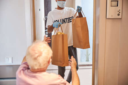Back view of a senior man reaching for paper bags in the hands of a young volunteerの写真素材