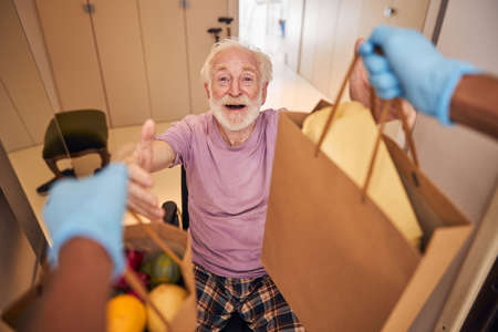 Pleased male pensioner receiving paper bags with alimentary products from the volunteer in latex...