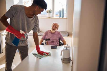 Smiling pleased disabled aged Caucasian person looking at the volunteer dusting the table in his presenceの写真素材