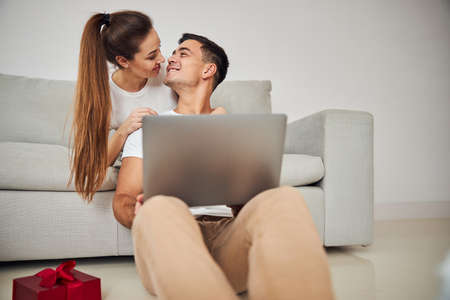 Handsome short-haired man with a laptop looking at his joyous wife with a long ponytail