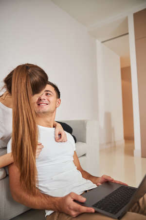 Cropped photo of gladsome young man smiling and looking at his girlfriend while holding laptop and...
