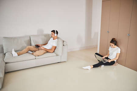 Focused young man and concentrated lady working on their laptops while sitting in different spots of...